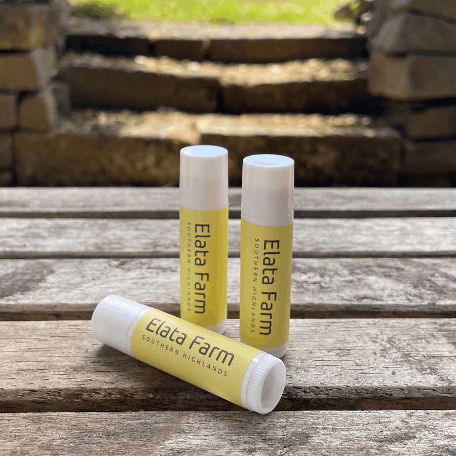 Three lip balm tubes from Elata Farm on a wooden surface with a stone wall background