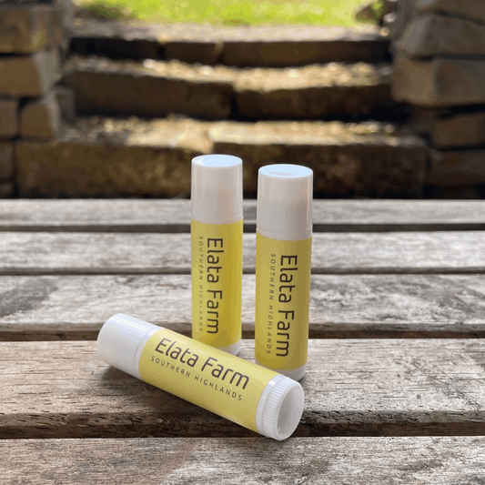Three lip balm tubes from Elata Farm on a wooden surface with a stone wall background